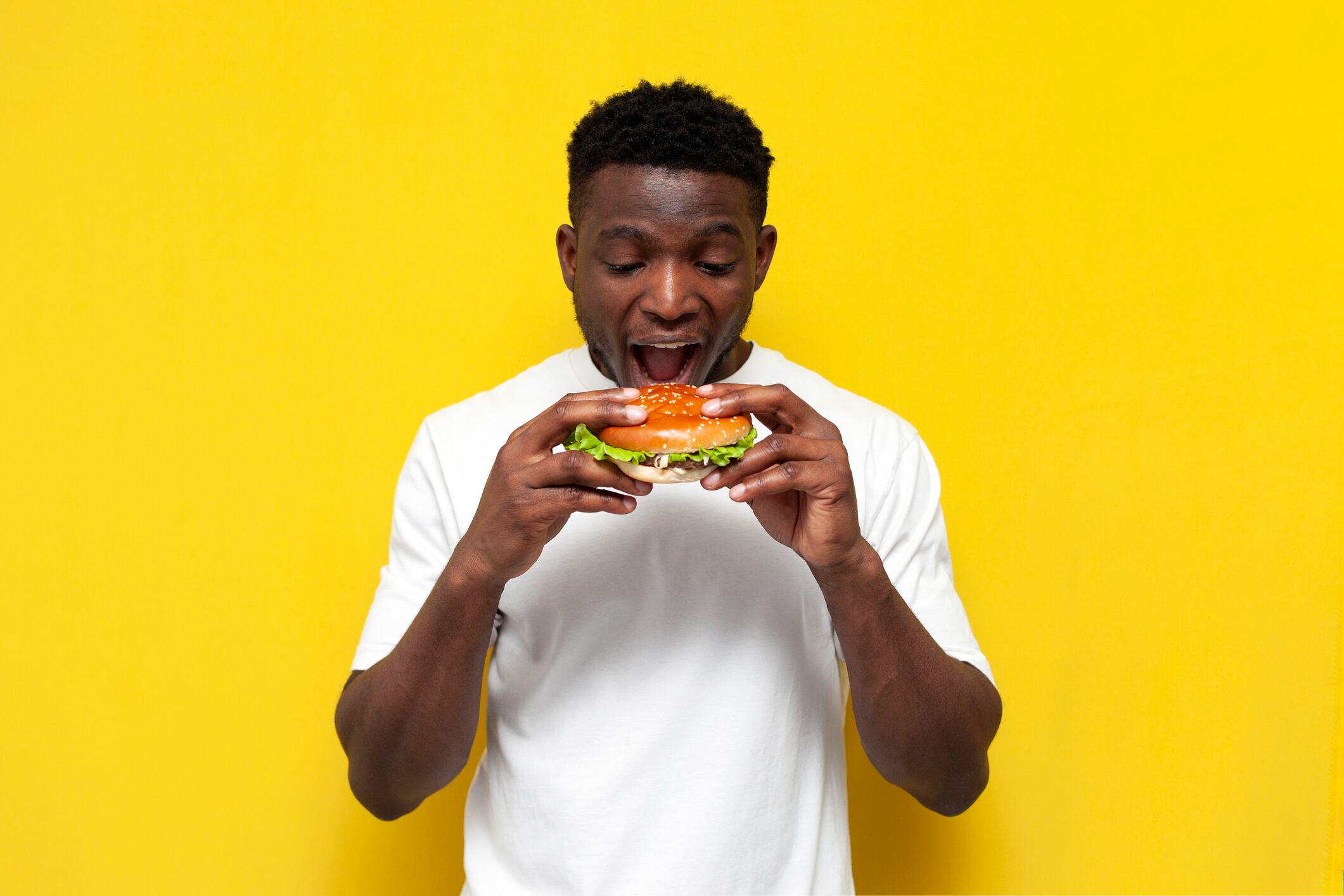 african american man in white t-shirt holding big burger and biting it, the guy eats fast food with his mouth open on yellow isolated background, the concept of unhealthy food and nutrition