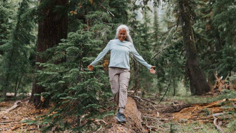 An older woman walking in the forest balancing on a fallen tree trunk
