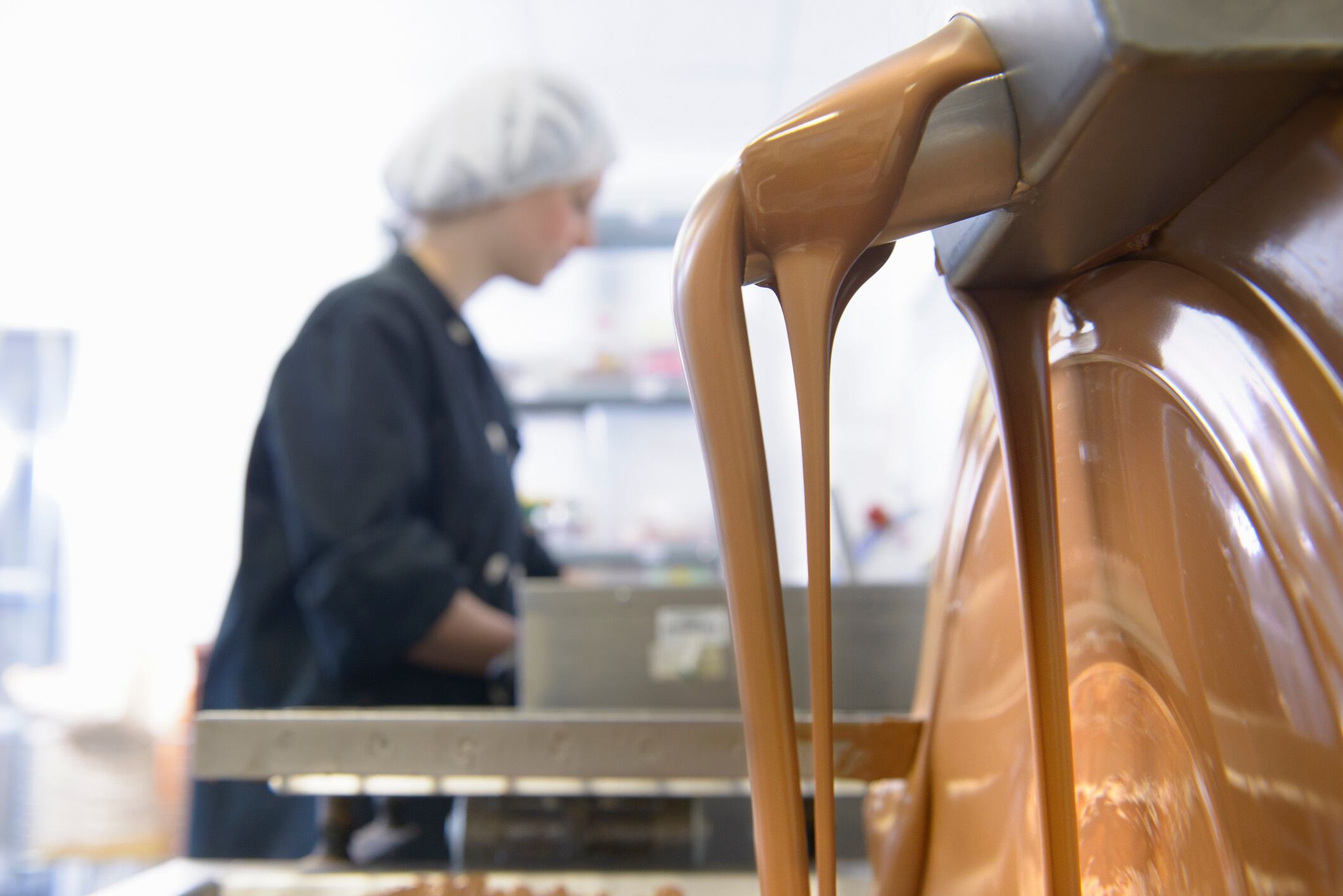 Chocolatier in background as chocolate pours from machine in chocolate factory