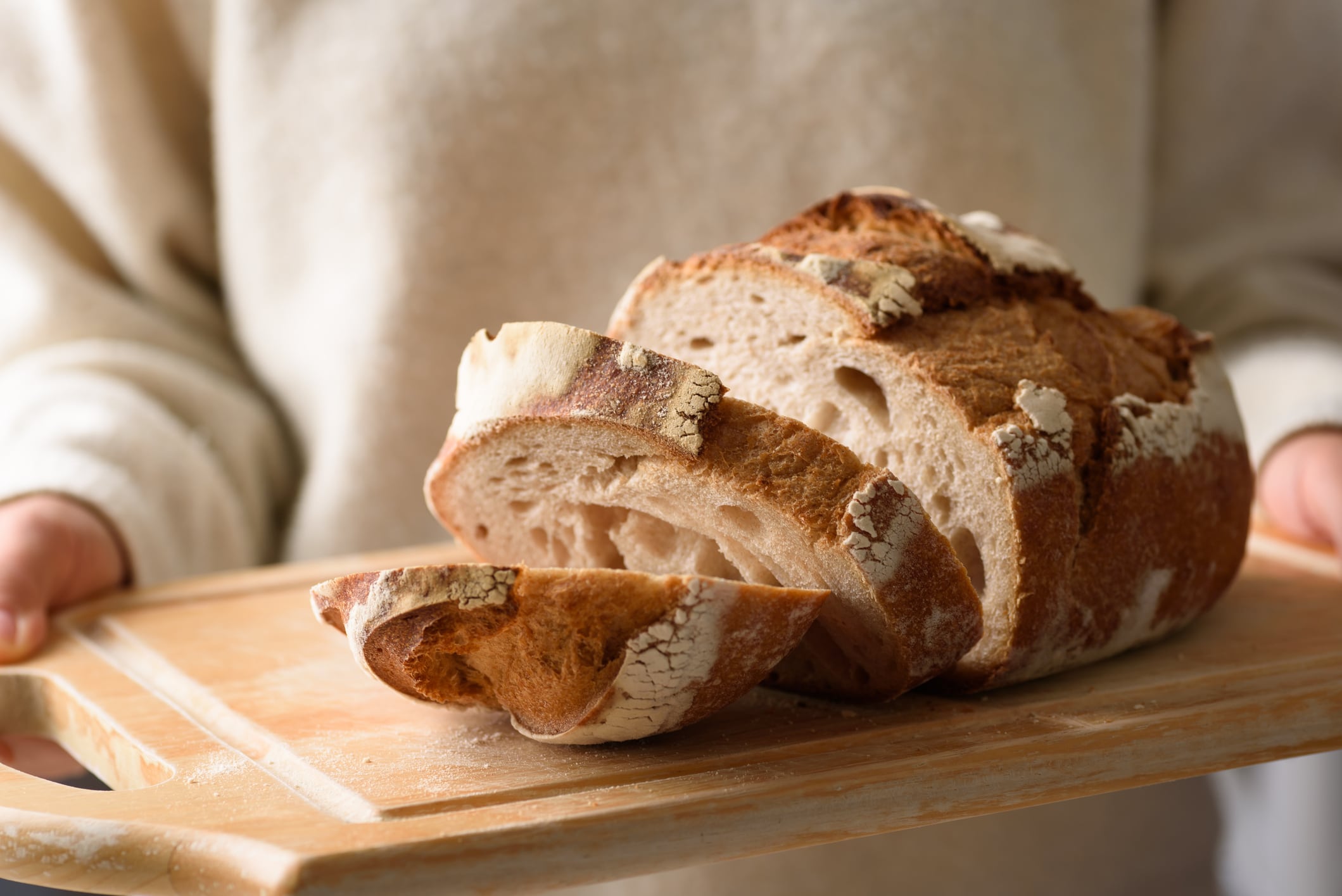 A close-up shot of a woman wearing a soft white long-sleeved shirt holding a slice of sourdough bread over a wooden cutting board